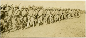 Horisontalt sepia -fotografi som viser rader med uniformerte soldater som marsjerer gjennom et gressfelt i formasjon mens de bærer rifler
